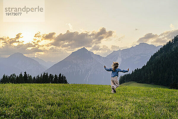 Mädchen läuft bei Sonnenuntergang auf Gras vor Bergen