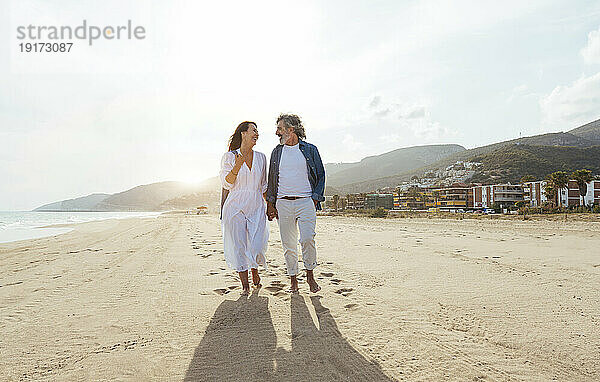 Glückliches Paar hält Händchen und spaziert an einem sonnigen Tag unter freiem Himmel am Strand