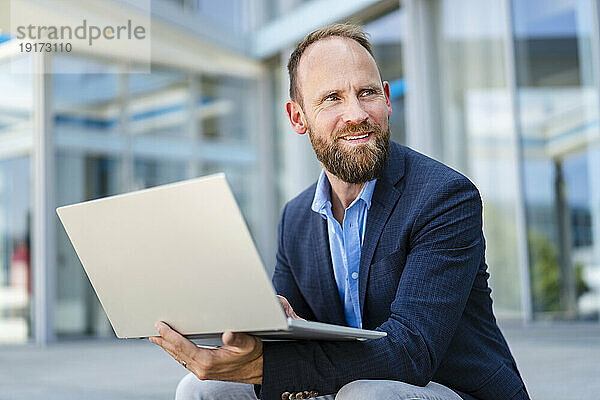 Geschäftsmann sitzt mit Laptop vor Bürogebäude