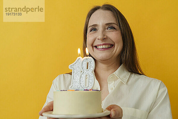 Glückliche Frau hält Geburtstagstorte vor gelbem Hintergrund