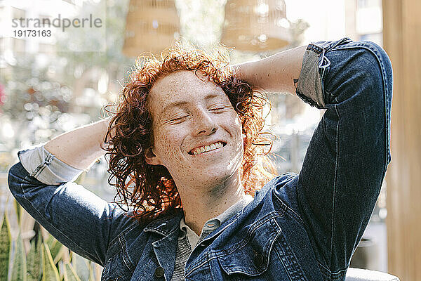 Happy redhead man sitting with hands behind head in cafe