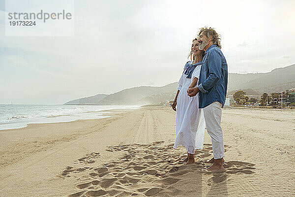 Lächelndes Paar hält Händchen und steht an einem sonnigen Tag am Strand