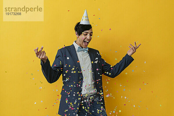 Joyful young man with party hat throwing confetti in studio