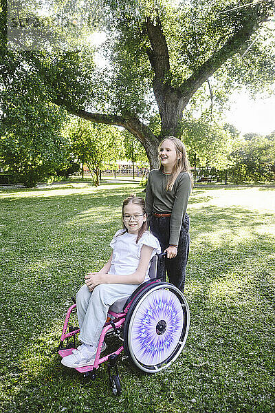 Girl sitting in wheelchair with friend standing behind at park