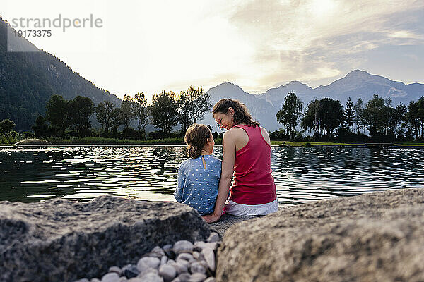 Mutter und Tochter sitzen bei Sonnenuntergang auf einem Felsen am See