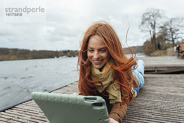 Lächelnde rothaarige Frau benutzt Tablet-PC am Pier am See
