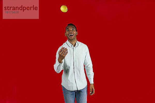 Fröhlicher Mann wirft Apfel vor rotem Hintergrund in die Luft