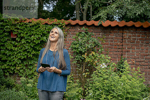 Happy mature woman standing with tablet PC in front of plants