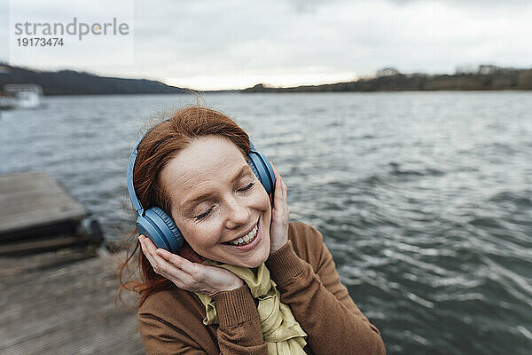Smiling woman listening to music through wireless headphones by lake