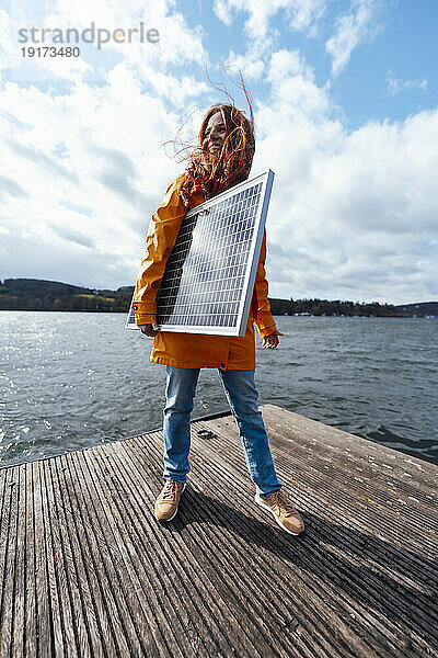 Frau mit Solarpanel steht auf Pier unter bewölktem Himmel am See