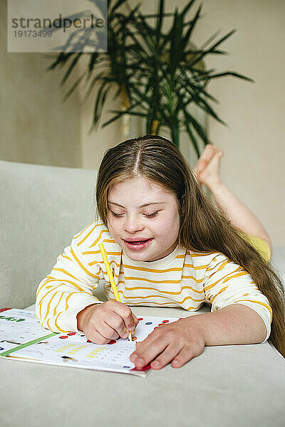 Lächelndes Mädchen mit Down-Syndrom  das zu Hause in ein Arbeitsbuch schreibt