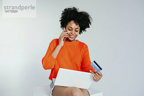 Lächelnde Frau mit Laptop und Kreditkarte  die auf dem Smartphone spricht