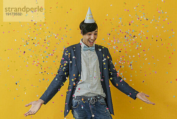 Glücklicher junger Mann mit Partyhut  der inmitten fallenden Konfettis genießt