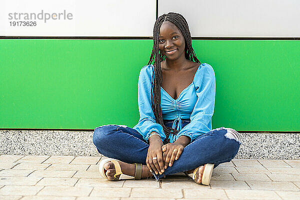 Lächelnde junge Frau sitzt mit gekreuzten Beinen vor der Wand