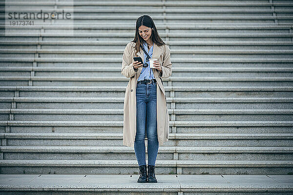 Glückliche Frau mit Kaffeetasse und Smartphone vor der Treppe