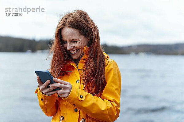 Happy redhead woman using smart phone in front of lake