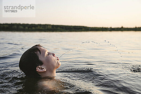 Junge spuckt bei Sonnenuntergang Wasser in den See