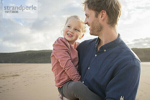 Smiling father and daughter at beach under cloudy sky