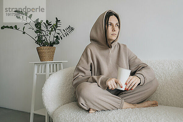 Nachdenkliche Frau sitzt mit Einwegbecher auf dem Sofa