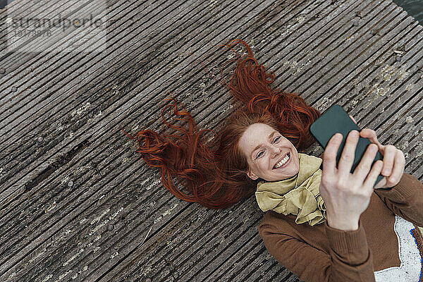 Happy redhead woman using mobile phone lying on pier