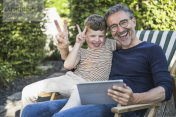 Fröhlicher Mann und Junge gestikulieren mit einem Tablet-PC und gestikulieren mit einem Friedenszeichen