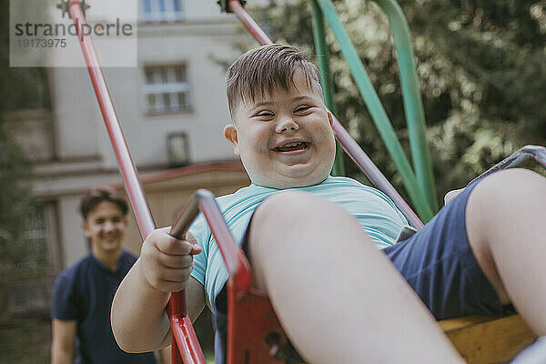 Glücklicher Junge mit Down-Syndrom  der mit seinem Bruder im Hintergrund auf der Schaukel spielt