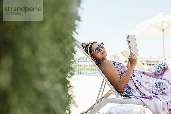 Glückliche Frau mit Sonnenbrille und entspannt mit einem Buch auf dem Liegestuhl