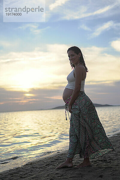 Schwangere Frau lächelt und steht unter bewölktem Himmel am Strand