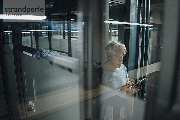 Leitender Geschäftsmann benutzt Mobiltelefon  gesehen durch Glas im Büro