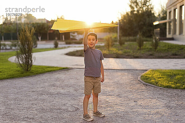 Lächelnder Junge hält Papierflieger in der Hand und steht im Park