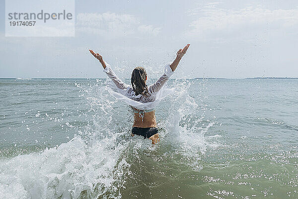 Frau mit erhobenen Armen steht und spritzt Wasser im Meer