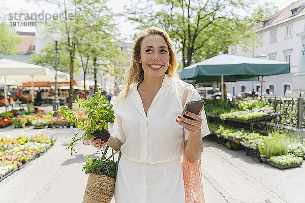 Glückliche Frau hält an einem sonnigen Tag Minzpflanze und Smartphone in der Hand