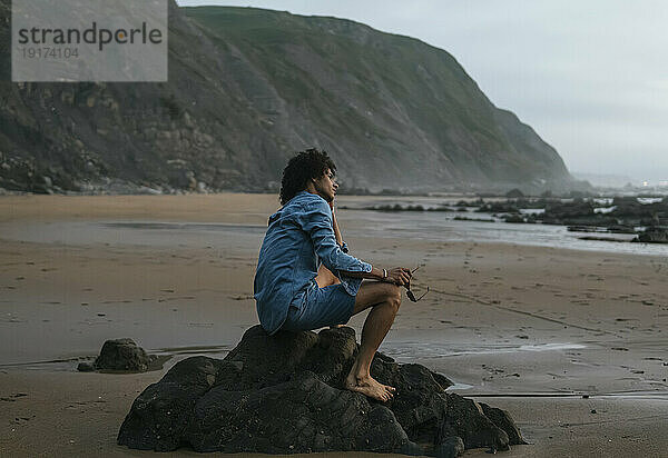 Junger Mann träumt davon  auf einem Felsen am Strand zu sitzen