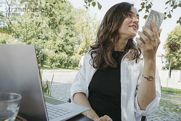 Lächelnde Geschäftsfrau  die am Tisch im Garten Voicemail per Smartphone aufzeichnet