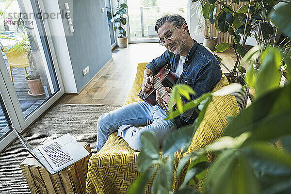 Mann spielt Gitarre und sitzt zu Hause auf dem Sofa neben dem Laptop