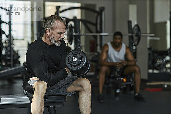 Reifer Mann trainiert mit Mann im Hintergrund im Fitnessstudio