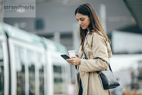 Junge Frau mit Kaffeetasse und Smartphone am Bahnhof