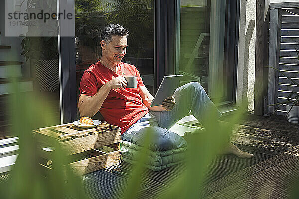 Mann entspannt sich auf Balkon mit digitalem Tablet und Tasse in der Hand