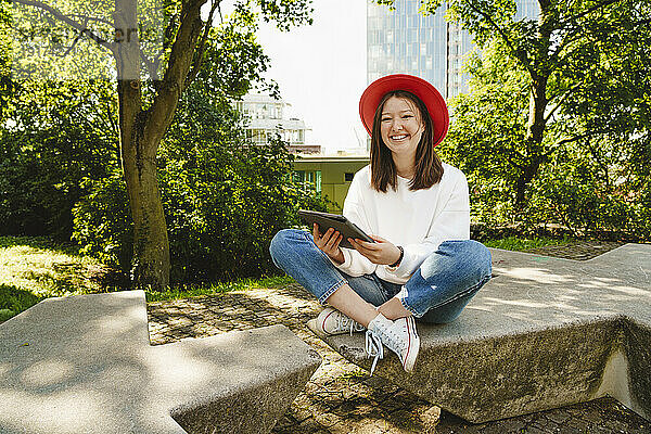 Glückliches Teenager-Mädchen mit Tablet-PC sitzt auf einer Bank im Park