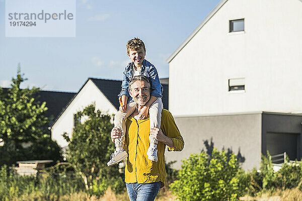 Smiling grandfather carrying grandson on shoulders in front of house