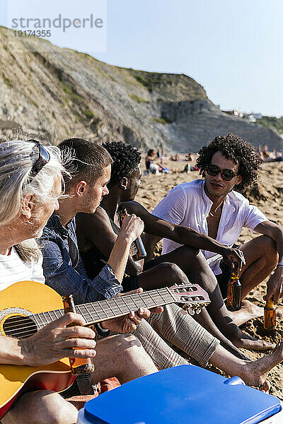 Reifer Mann mit Gitarre sitzt neben Freunden und trinkt Bier am Strand