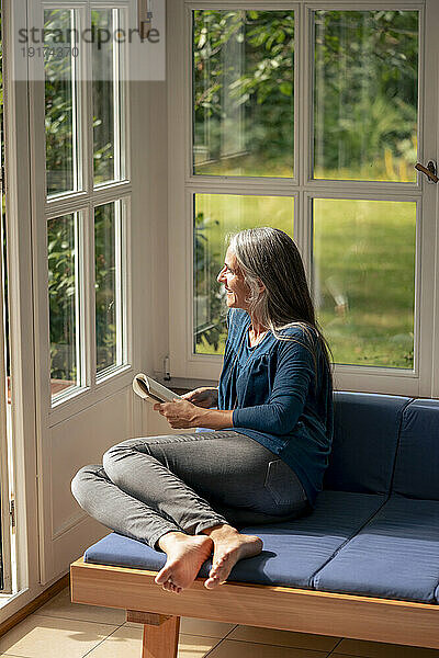Lächelnde Frau  die zu Hause mit einem Buch auf dem Sofa sitzt