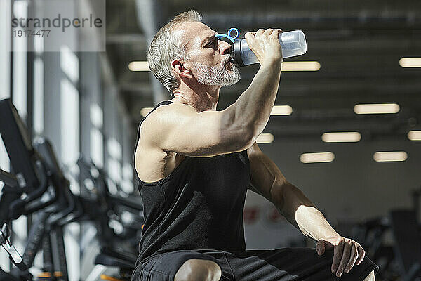 Reifer Mann trinkt Wasser im Sonnenlicht im Fitnessstudio