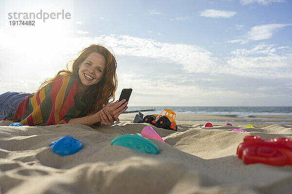 Glückliche Frau mit Smartphone  die am Strand im Sand liegt