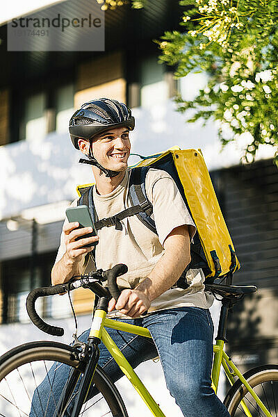 Glücklicher junger Zusteller mit Smartphone auf dem Fahrrad
