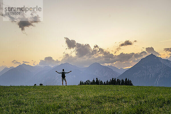 Hiker with arms outstretched standing on grass in front of mountains