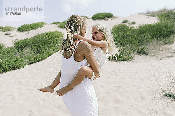 Glückliche Mutter trägt ihre Tochter an einem sonnigen Tag am Strand