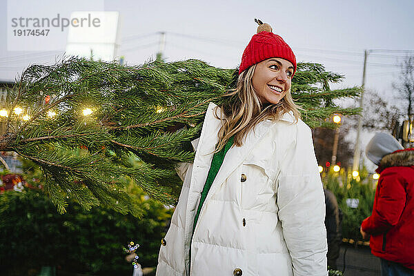 Nachdenkliche Frau mit Strickmütze trägt Weihnachtsbaum auf dem Markt