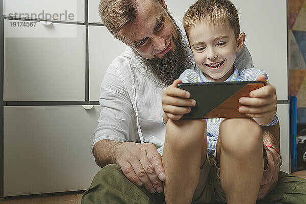 Smiling boy sitting on father's lap and using smart phone at home