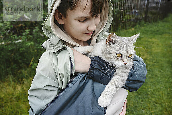 Smiling boy wearing jacket carrying cat in garden